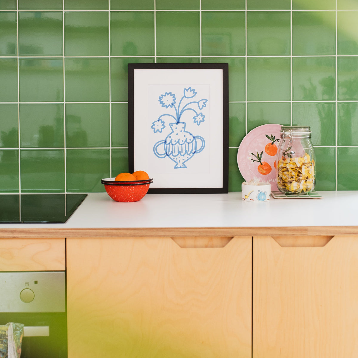 A framed art print with a blue floral vase design hangs on a kitchen counter against a green tiled backsplash. The scene includes light wood cabinets, a bowl of oranges, and a glass jar of snacks.
